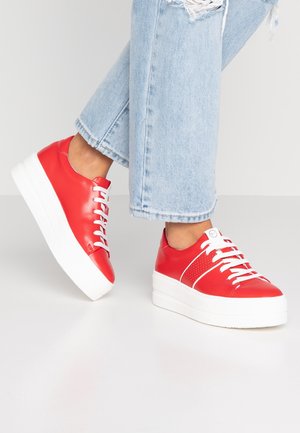 Osoba nosząca czerwone sneakersy na platformie z białymi sznurowadłami oraz jasnoniebieskie rozdarcia dżinsowe, stojąca na białej podłodze.