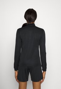 Femme à la peau foncée et aux longs cheveux foncés attachés, portant un haut à manches longues noir et un short noir, vue de dos sur un fond blanc.