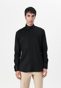 Μαύρο πουκάμισο με μακριά μανίκια και ψηλό γιακά, με κουμπιά, και λεία, ματ υφή. Φορεμένο με ανοιχτό μπεζ παντελόνι.