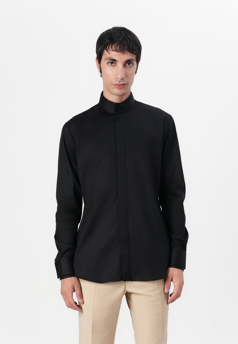 Μαύρο πουκάμισο με μακριά μανίκια και ψηλό γιακά, με κουμπιά, και λεία, ματ υφή. Φορεμένο με ανοιχτό μπεζ παντελόνι.