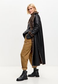 Mujer con cabello corto rubio, vistiendo un largo abrigo negro con borde de piel de leopardo, suéter gris, pantalones beige y botas negras de cordones, de pie de lado.