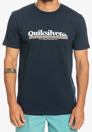 Mand iført en mørkeblå Quiksilver T-shirt med hvid og orange logotekst og lyseblå shorts, viser en tatovering på venstre underarm.