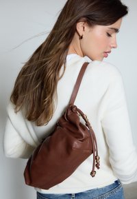 Borsa a tracolla in pelle marrone con design arricciato e dettagli in metallo dorato, indossata sulla spalla sopra un maglione bianco.