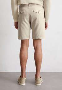 Shorts en coton beige avec une coupe ajustée, dotés de deux poches arrière avec fermetures à bouton et d'un devant plat, sur un fond neutre.