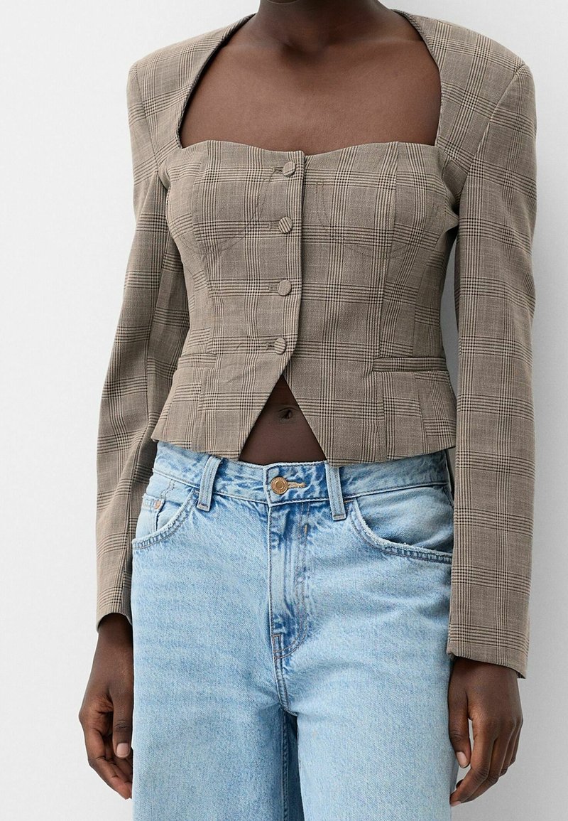 Femme portant un blazer à carreaux beige à col carré avec boutons et un jean taille haute bleu clair, debout devant un fond uni.