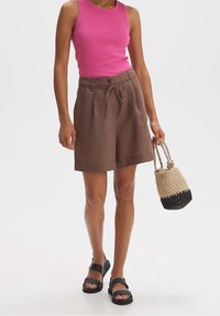 Shorts en lin marron avec une taille nouée, assortis à un débardeur côtelé rose et des sandales noires, accompagnés d'un sac tissé au design dégradé.