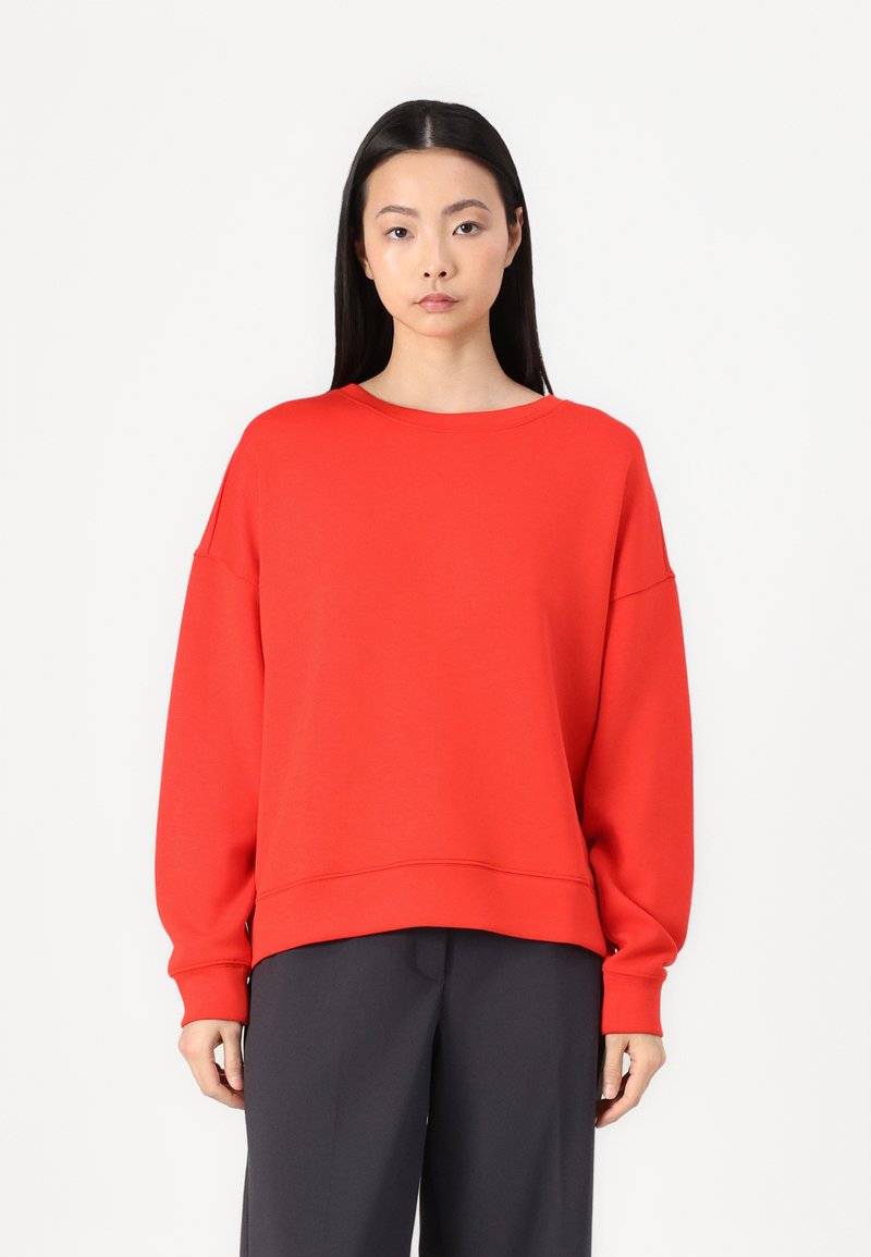 Felpa rossa oversize con scollo rotondo e maniche lunghe, caratterizzata da una texture morbida e una vestibilità rilassata, abbinata a pantaloni scuri.