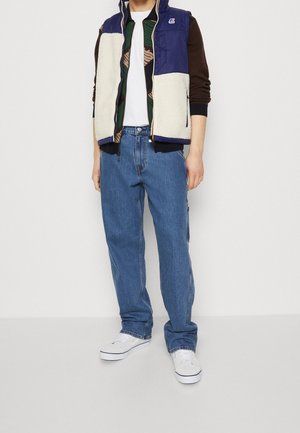 Person trägt eine blau-beige ärmellose Weste über einer gemusterten Jacke, blaue Jeans und weiße Sneakers, steht vor einem einfarbigen weißen Hintergrund.