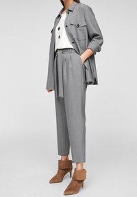 Vrouw in lichtgrijze broek met riem en bijpassende lange overshirt met opgerolde mouwen, witte top, gelaagde kettingen en bruine enkellaarsjes.
