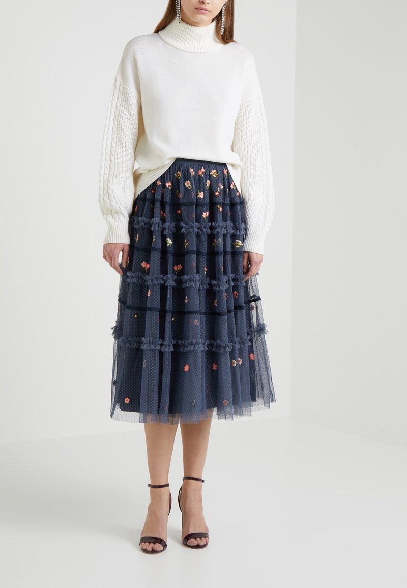 Femme portant un pull à col roulé en maille torsadée blanche et une jupe midi à volants bleu marine avec broderies florales et superposition en voile transparent.
