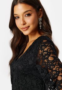 Černý krajkový top s výstřihem do V a dlouhými rukávy, zdobený květinovými vzory a texturovaným povrchem. Doplněno třpytivým náušnicemi.