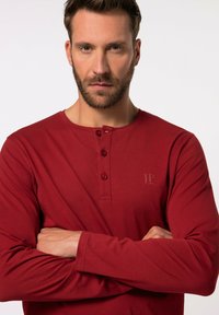 Maglietta a maniche lunghe rossa in stile henley realizzata in tessuto morbido, con un colletto a tre bottoni, texture a coste e un discreto dettaglio del logo sul petto.