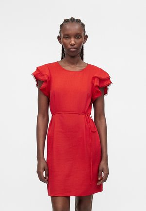 Donna con capelli intrecciati che indossa un vestito corto rosso con maniche a volant stratificati e una sottile cintura in vita, in piedi davanti a uno sfondo semplice.