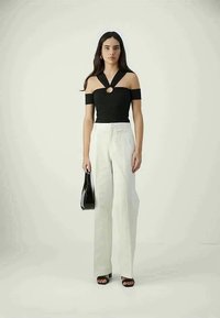 Frau steht in einem schwarzen schulterfreien Oberteil mit einem Frontring-Detail, weißen weitem Hosen und schwarzen Sandalen und hält eine schwarze Handtasche.