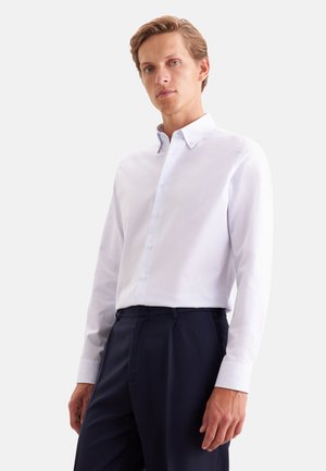 Jeune homme portant une chemise blanche à boutons et un pantalon bleu marine foncé sur un fond clair uni.