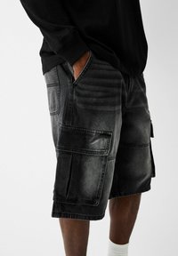 Person med svarta cargo-shorts med stora sidofickor och en svart långärmad skjorta, hand i fickan, som står mot en vit bakgrund.