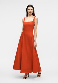 Vrouw staand in een mouwloze, vierkante hals, oranje jurk tot op de vloer met zwarte, gespen hoge hakken op een effen witte achtergrond.