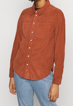 Person iført en rustfarvet kordfløjls-skjorte med knapper og to brystlommer samt lyseblå cowboybukser mod en ensfarvet baggrund.