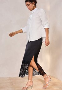 Žena v bílé košili na knoflíky a černé sukni s krajkovým lemem, která kráčí, obutá ve stříbrných sandálech na podpatku s pásky, na neutrálním pozadí.