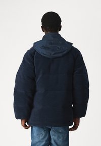 Navy blå pufferjakke med en teksturert, fleece-overflate og en kontrastfarget nylonhette. Har en boksformet silhuett og vide ermer.