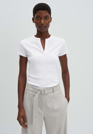 Frau mit kurzen Haaren, die ein weißes Kurzarm-Top und hellbeige, mit Gürtel versehene Hosen trägt, steht mit einer Hand in der Tasche.