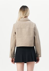 Cropped beige wool jacket with a collar, visible seams, and snap buttons; worn over a black pleated skirt, showcasing the jacket's back.