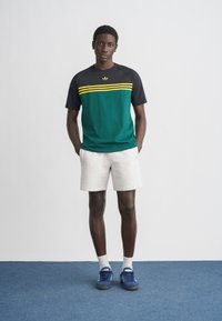 T-shirt de manga curta verde e preta com riscas amarelas; combinado com calções brancos e ténis azuis. Design simples, ajuste casual.