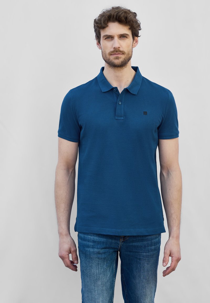 Blå kortärmad polo med krage och tvåknapplikning, tillverkad av strukturerad tyg, tillsammans med urblekta blå jeans.