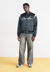 Svart sateng bomberjakke med hvite detaljer og "Jordan"-bokstaver, sammen med vide, falmede grå jeans og svart-hvite sneakers.