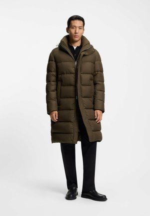 Olijfgroene pufferjas met een lange snit, voorzien van gestikte panelen, ritssluiting, een verstelbare capuchon en zijzakken. Draag het over een zwarte broek.