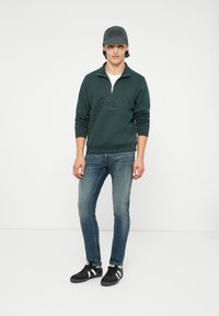 Młody mężczyzna ubrany w ciemnozieloną bluzę z suwakiem do ćwiartki, niebieskie dżinsy, czapkę i czarne trampki w białe paski, stojący na białym tle.