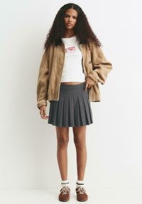 Une jeune femme aux cheveux bouclés porte une veste en velours côtelé beige, un débardeur blanc avec un texte rouge, une jupe plissée grise, des chaussettes blanches et des chaussures marron.