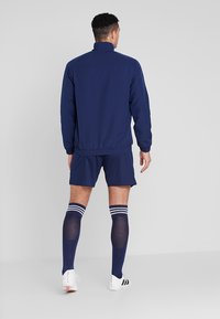 Marinefarbene Sportjacke und Shorts mit elastischem Bund; kombiniert mit kniehohen Socken mit weißen Streifen.