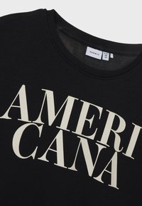 Sweatshirt noir avec un grand texte "AMERICANA" de couleur crème sur le devant. Encolure côtelée et texture douce. Étiquette de marque visible.