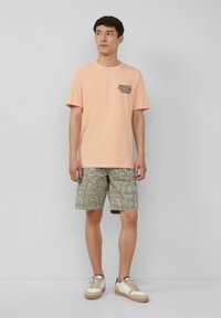 Licht perzik kleurige katoenen t-shirt met een bedrukte logo op de borst, gecombineerd met groene gestreepte shorts en tan sneakers met witte accenten.