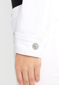 Valkoinen farkkutakin hiha, jossa hopeinen painonappi, esittelee sileän tekstuurin ja siistit ompeleen yksityiskohdat.