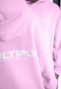 Svetloružová mikina s kapucňou vyrobená z mäkkej látky, s veľkým bielym textom "MULTIPLY" a menším textom "MULTIPLY APPAREL CLUB" na chrbte.