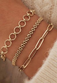 Drei goldene Armbänder an einem Handgelenk: eine verbundene ovale Kette, eine texturierte quadratische Gliederkette und eine glatte längliche Gliederkette, die unterschiedliche Designs präsentieren.