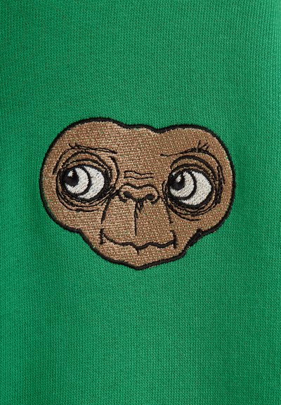 Zelená látka s textúrovanou vyšívanou hnedou tvárou mimozemšťana so veľkými očami a zaobleným tvarom, umiestnená v strede odevu.