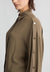 Blusa verde oliva con colletto, maniche lunghe e dettagli con bottoni sulle spalle. Realizzata in un tessuto leggero e morbido con una silhouette elegante.