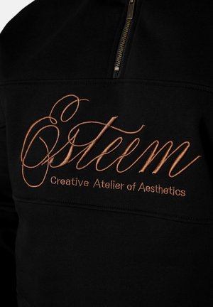 Schwarzer Stoff mit kupferfarbener Stickerei mit der Aufschrift "Östeem Creative Atelier of Aesthetics" unterhalb eines teilweisen Reißverschlusses.