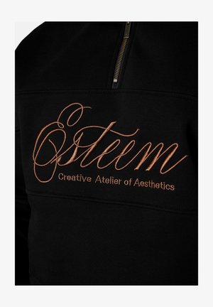 Schwarzer Stoff mit kupferfarbener Stickerei mit der Aufschrift "Östeem Creative Atelier of Aesthetics" unterhalb eines teilweisen Reißverschlusses.