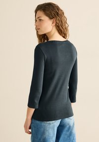 Černý dlouhý rukáv top z jemné tkaniny s oblečeným kulatým výstřihem, s přiléhavými rukávy a mírně zakřiveným lemem. V kombinaci se světlými džínami.