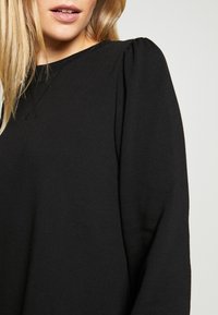 Pull noir avec un col rond, un tissu texturé et des manches bouffantes, présentant une apparence lisse et douce.
