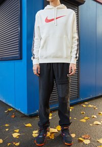 Vit Nike hoodie med röd logotyp, svarta sweatpants med texturerad lagning och svarta sneakers med orange detaljer, mot en blå bakgrund.