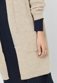 Béžový pletený cardigan s texturovaným povrchem, který má velkou přední kapsu. Nosený přes námořnické šaty s malým geometrickým vzorem.
