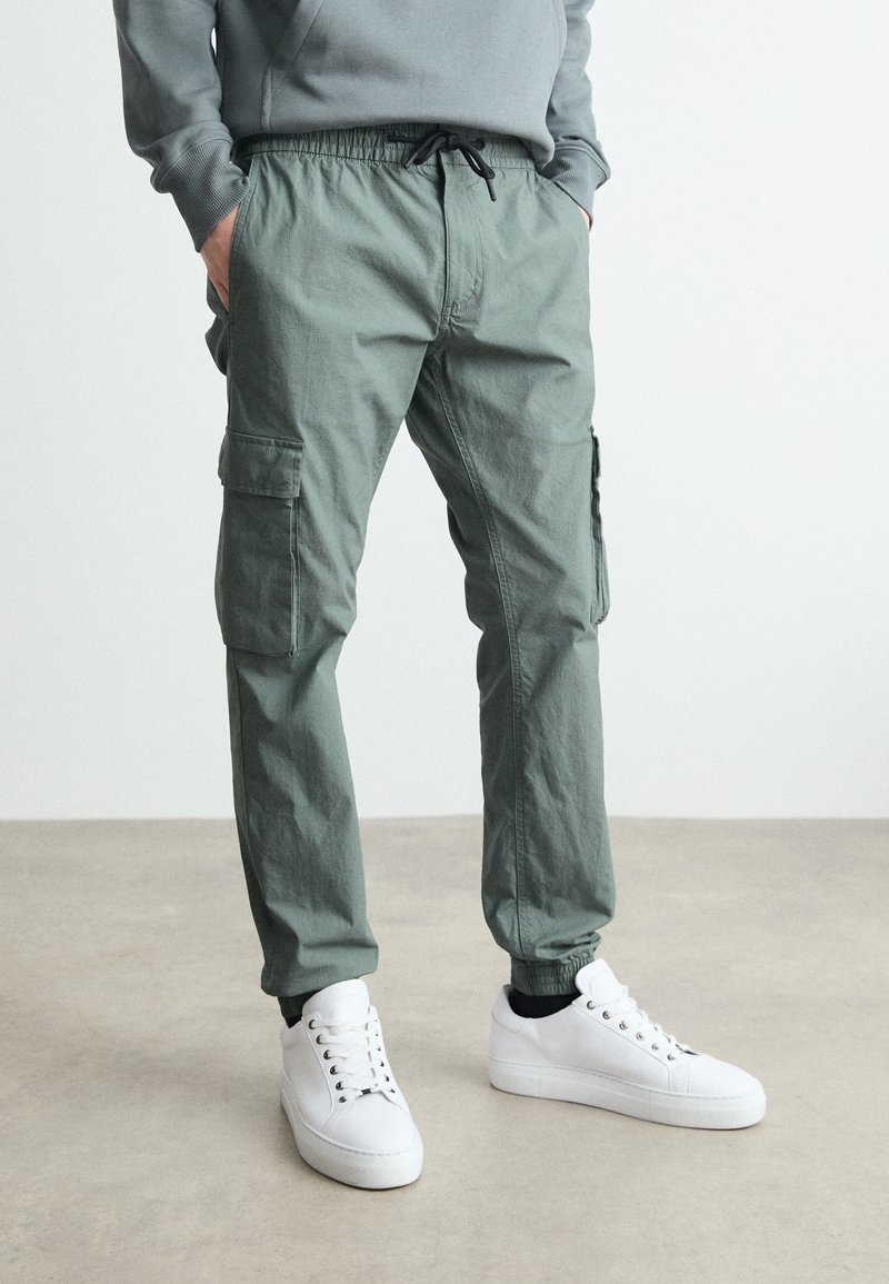 Zielone bawełniane spodnie typu cargo z elastycznym pasem, bocznymi kieszeniami i zwężanymi nogawkami, połączone z białymi trampkami. Prosty, praktyczny design.