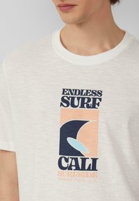 Witte t-shirt met korte mouwen met een blauw en perzikkleurig grafisch ontwerp met de tekst "ENDLESS SURF," "CALI," en "VENICE PIER, L.A." gedrukt.