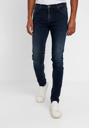 Hombre vestido con vaqueros ajustados azul oscuro, camiseta blanca y zapatillas blancas, de pie frente a un fondo claro y liso.