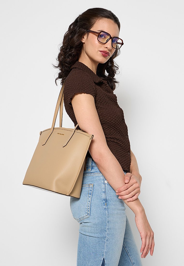 Borsa tote beige in finta pelle liscia, design strutturato, apertura superiore, doppi manici. Modella che indossa jeans azzurri e top marrone.